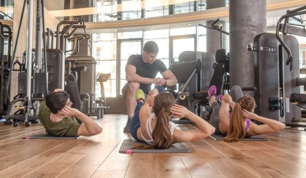 Fitness instructor timing three young people doing lateral crunch exercise with raised legs for abdominal muscles indoors at the gym